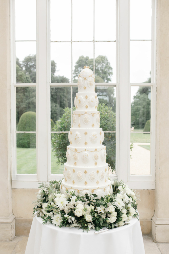 By Yevnig luxury 7-tier wedding cake in ivory white and gold sits atop a table in the Great Conservatory at Syon Park which has been decorated with fresh florals and foliage in white and gree by Emma Soulsby