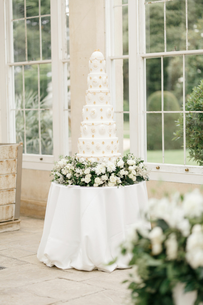 By Yevnig luxury 7-tier wedding cake in ivory white and gold sits atop a table in the Great Conservatory at Syon Park which has been decorated with fresh florals and foliage in white and gree by Emma Soulsby
