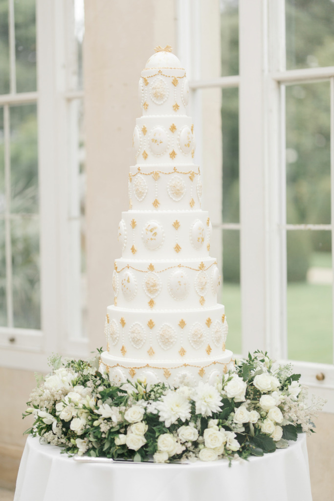 By Yevnig luxury 7-tier wedding cake in ivory white and gold sits atop a table in the Great Conservatory at Syon Park which has been decorated with fresh florals and foliage in white and gree by Emma Soulsby