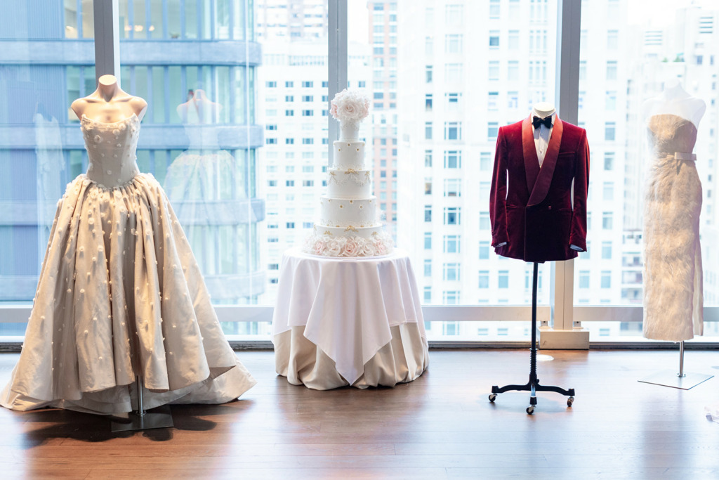 Image of luxury wedding cake By Yevnig next to bridal dresses from Suzie Turner Couture and a red velvet tux jacket from Hunstaman Savile Row against the backdrop of Manhattan high rise buildings.