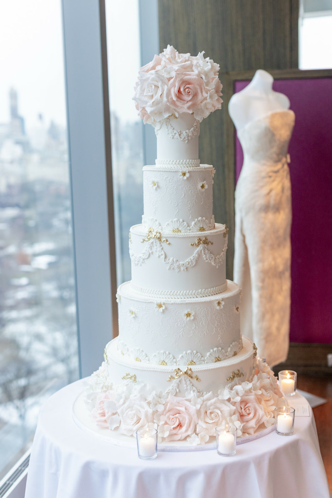 Luxury 5-tier wedding cake, By Yevnig, decorated with hand-crafted sugar roses, and delicate piped decorations stands on top of a table by a window at the Mandarin Oriental New York. In the background a Suzie Turner Couture wedding dress is displayed on a mannequin