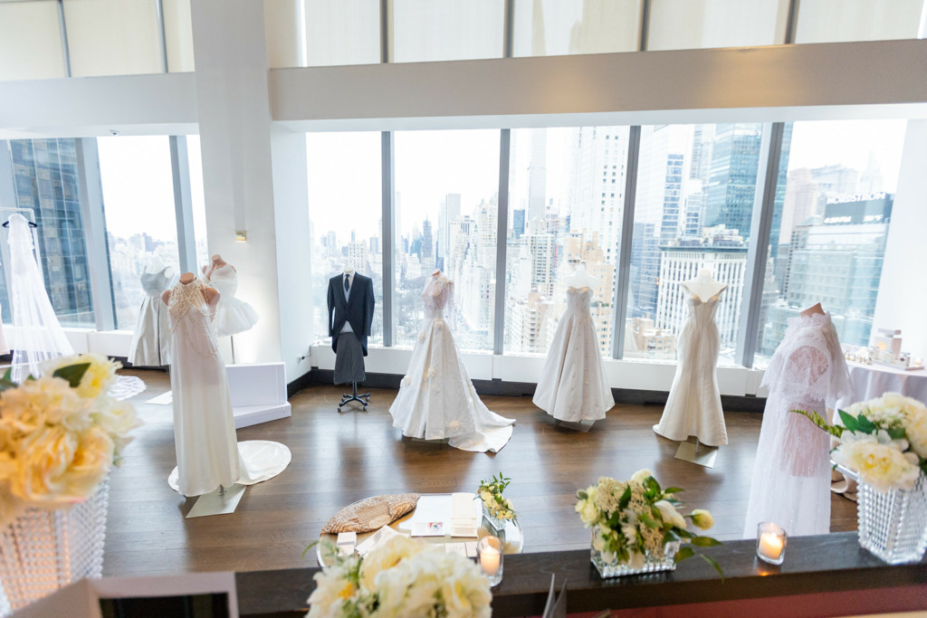 Selection of Suzie Turner Couture gowns on manquins at the Mandarin Oriental, New York, in front of a window with high rise Manhattan buildings.