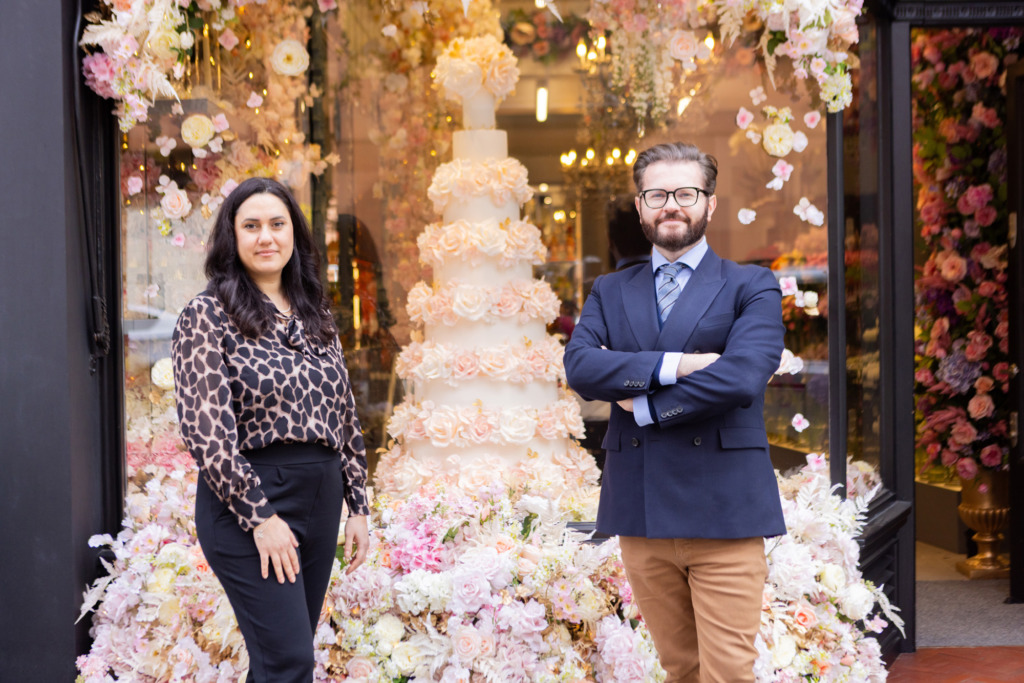 Yevnig Davis and Neill Strain stand outside Neill Strain Floral Couture, Mayfair London. Behind the florist shop window is surrounded by fresh floral installation with a By Yevnig floral wedding cake inside the window.